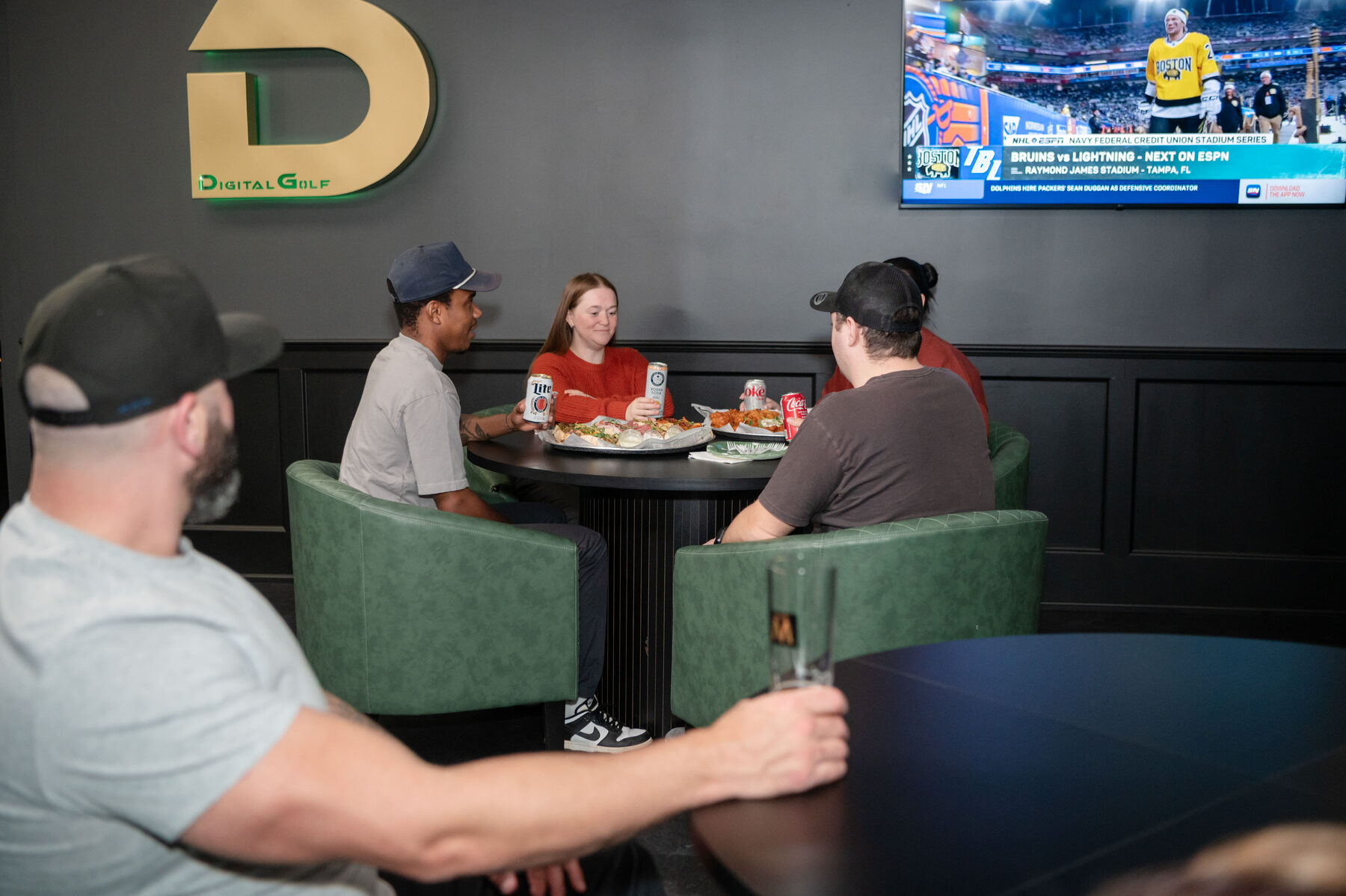 Customers relaxing with food and drinks in the lounge area under the Digital Golf logo, with sports playing on the TV.